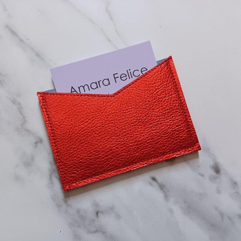 May include: A bright red leather card holder with the text "Amara Felice" on a light purple card. The card holder is rectangular with a unique angled top design. The card holder is made of textured leather.