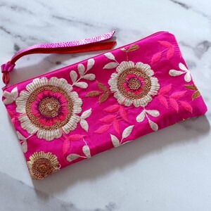 Op de afbeelding: Een felroze clutch met ingewikkelde bloemenborduurwerk. Het ontwerp heeft grote, geborduurde bloemen met gouden en roze accenten en witte bladeren. Er is een bijpassende roze band bevestigd. De clutch is ongeveer 25 cm lang.
