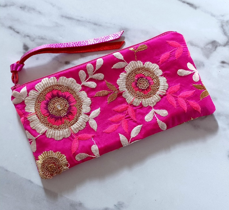 Op de afbeelding: Een felroze clutch met ingewikkelde bloemenborduurwerk. Het ontwerp heeft grote, geborduurde bloemen met gouden en roze accenten en witte bladeren. Er is een bijpassende roze band bevestigd. De clutch is ongeveer 25 cm lang.
