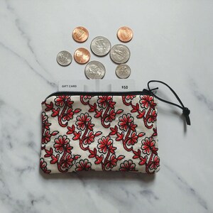 Op de afbeelding: Een kleine, cr&egrave;mekleurige etui met ritssluiting, een rood bloemmotief en een zwarte ritstrekker. Boven de etui liggen verschillende munten en een cadeaukaart. Op de cadeaukaart staat "$50".