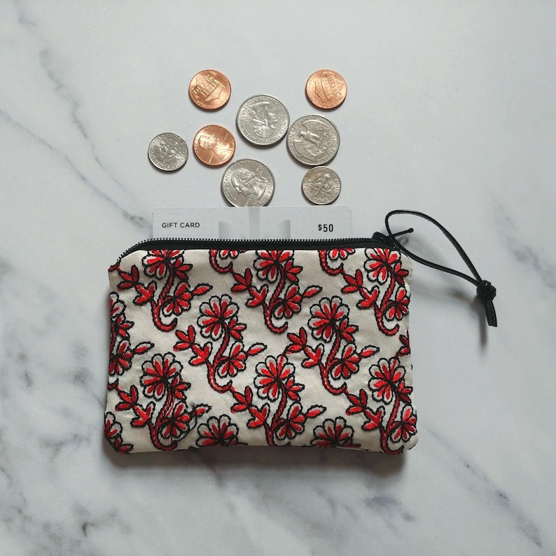 Op de afbeelding: Een kleine, cr&egrave;mekleurige etui met ritssluiting, een rood bloemmotief en een zwarte ritstrekker. Boven de etui liggen verschillende munten en een cadeaukaart. Op de cadeaukaart staat "$50".