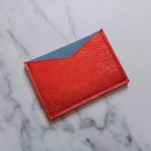 May include: A rectangular card holder with a vibrant red exterior and a light blue interior. The card holder is made of textured material and has a stitched border. The design features a unique angled top edge.