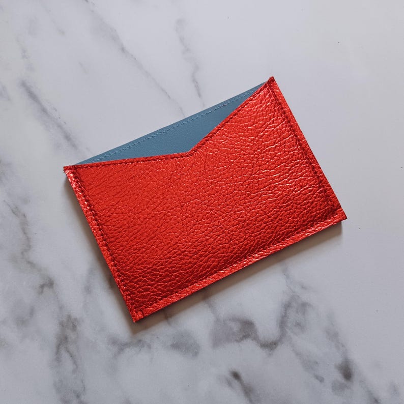 May include: A rectangular card holder with a vibrant red exterior and a light blue interior. The card holder is made of textured material and has a stitched border. The design features a unique angled top edge.