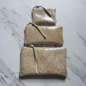 May include: Three beige, rectangular zippered pouches of varying sizes are stacked on top of each other. Each pouch has a matching beige wrist strap attached to the zipper. The pouches have a textured, speckled pattern.