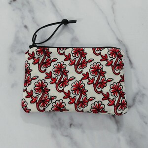 Op de afbeelding: Een witte etui met ritssluiting en een herhalend bloemmotief in rood en zwart. De etui heeft een zwart koord aan de rits. Het bloemmotief heeft rode bloemen en bladeren.