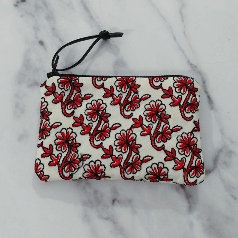 Op de afbeelding: Een witte etui met ritssluiting en een herhalend bloemmotief in rood en zwart. De etui heeft een zwart koord aan de rits. Het bloemmotief heeft rode bloemen en bladeren.