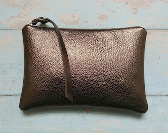 May include: A rectangular metallic bronze clutch bag with a pebbled texture. The bag features a top zipper closure with a long, thin matching leather pull tab attached to the zipper slider, providing a simple and functional design for carrying items.