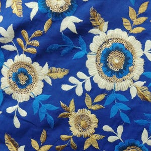 Op de afbeelding: Close-up van een koningsblauwe stof met geborduurde bloempatronen. Het ontwerp toont witte, gouden en blauwe bloemen met ingewikkelde details. De bladeren zijn in tinten goud, wit en blauw.