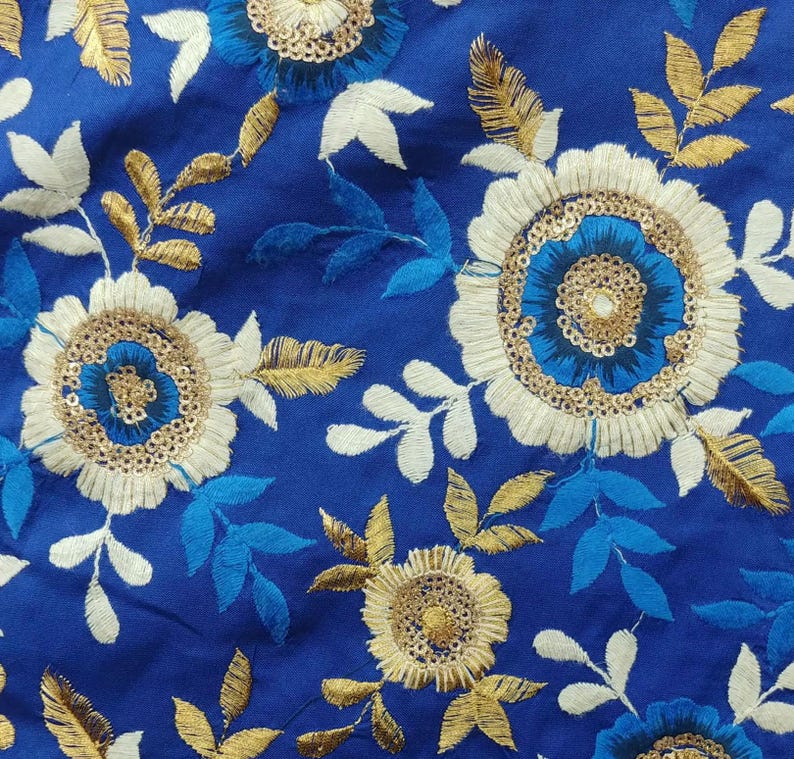 Op de afbeelding: Close-up van een koningsblauwe stof met geborduurde bloempatronen. Het ontwerp toont witte, gouden en blauwe bloemen met ingewikkelde details. De bladeren zijn in tinten goud, wit en blauw.