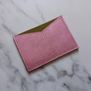 May include: A pink, textured leather card holder with an olive green interior. The card holder is rectangular with a v-shaped opening. The item is set against a marble background.