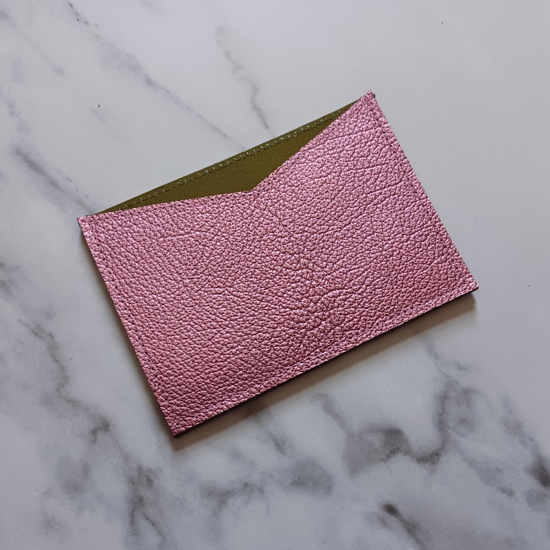 May include: A pink, textured leather card holder with an olive green interior. The card holder is rectangular with a v-shaped opening. The item is set against a marble background.