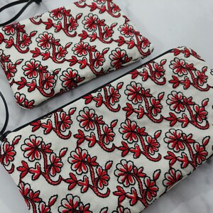 Op de afbeelding: Twee witte stoffen etuis met een rood bloemenborduurontwerp. De borduursels hebben rode bloemen en bladeren met zwarte contouren. Elke etui heeft een zwarte rits en een zwarte band.