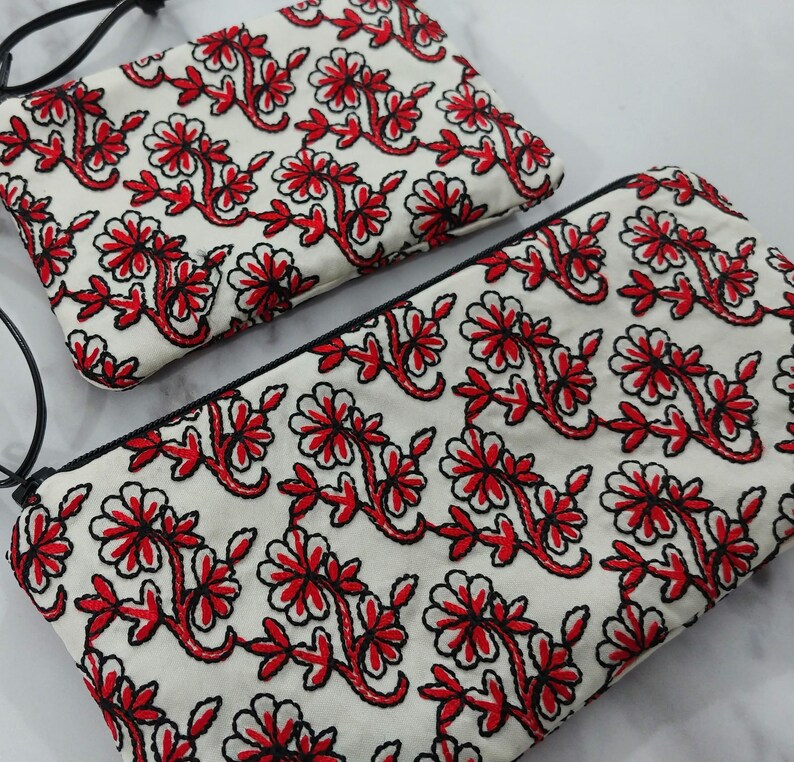 Op de afbeelding: Twee witte stoffen etuis met een rood bloemenborduurontwerp. De borduursels hebben rode bloemen en bladeren met zwarte contouren. Elke etui heeft een zwarte rits en een zwarte band.