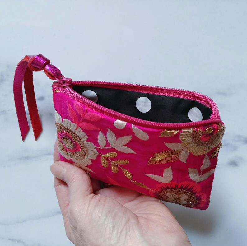 Op de afbeelding: Een felroze geborduurd etui met ritssluiting en leren trekker. De tas heeft gouden en witte bloemenborduursels op de voorkant. De binnenkant is zwart met witte stippen. Een klein, stijlvol accessoire.