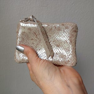 May include: A small, rectangular, light-colored coin purse with a textured, snakeskin-like pattern. The purse has a zipper closure and a matching wrist strap. The color is a mix of beige, silver, and pink.