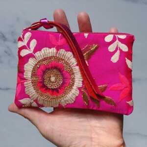 Op de afbeelding: Een felroze geborduurde clutch met een bloemmotief. De clutch heeft een grote, gedetailleerde bloem in het midden met gouden en cr&egrave;mekleurige accenten. Een rode band en rits maken het ontwerp compleet. De clutch wordt in een hand gehouden.