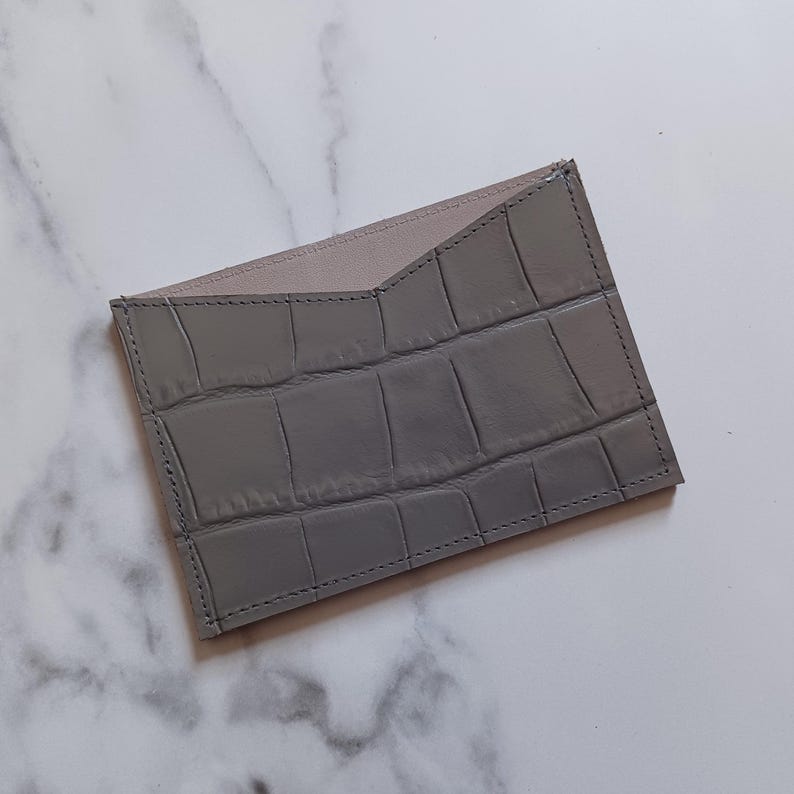 May include: A gray cardholder with a crocodile skin texture. The interior is a light beige color. The cardholder is rectangular with a stitched border.