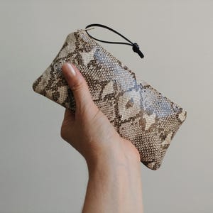May include: A rectangular clutch with a snakeskin pattern in shades of beige, brown, and gray. The clutch has a black cord loop attached to one end. The bag is held in a hand.
