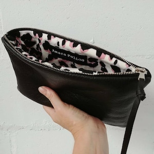 May include: Black leather clutch with a zipper closure and a pink and black leopard print lining. The clutch has a small strap and the words "Amara Felice" are printed on the lining.