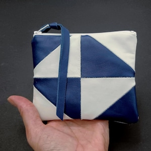 May include: A rectangular pouch featuring a geometric patchwork design of alternating navy blue and off-white leather panels. The pouch includes a matching navy blue leather wrist strap attached to the top zipper and white stitching.