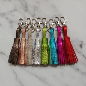 May include: A collection of eight leather tassels in various metallic and textured finishes, including bronze, copper, beige, silver, lime green, turquoise, magenta, and red. Each tassel features a silver-toned metal lobster claw clasp.