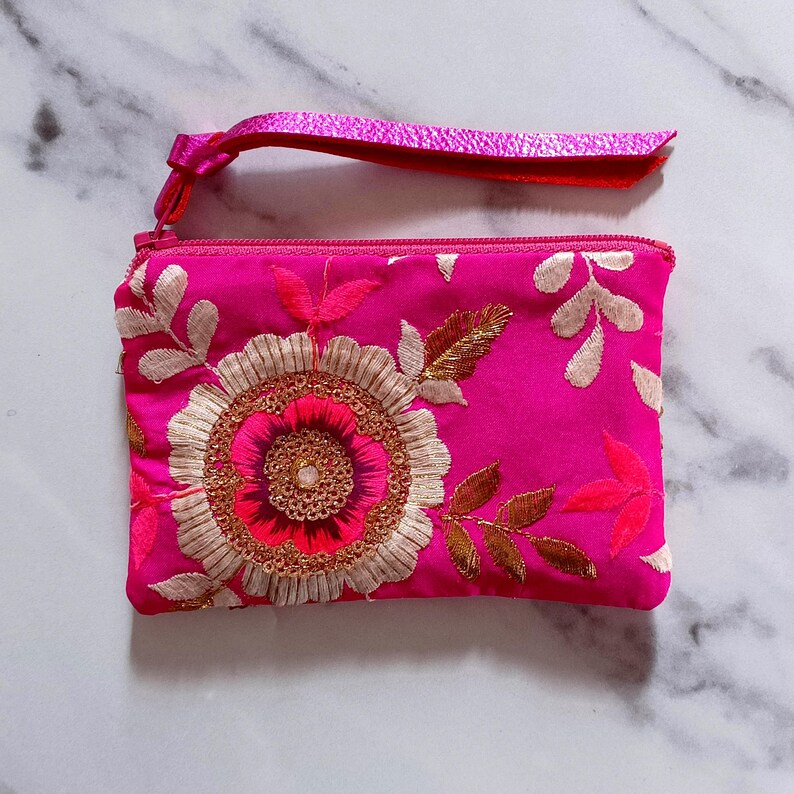 Op de afbeelding: Een felroze clutch met een polsbandje. De clutch heeft een geborduurd bloemmotief met goud, wit en roze draad. De rits en het polsbandje zijn in bijpassende roze kleur.