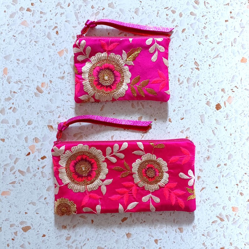 Op de afbeelding: Twee felroze geborduurde ritszakjes met bloemmotieven. De zakjes hebben een polsbandje en zijn versierd met gouden en witte draad. Het bloemmotief staat centraal en heeft een gedetailleerd, gestructureerd uiterlijk.