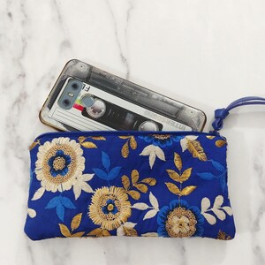 Op de afbeelding: Een koningsblauwe geborduurde clutch met een polsbandje, versierd met gouden, witte en blauwe bloemmotieven. Een smartphone met een cassettebandje hoesje ligt erin, een modieus accessoire.