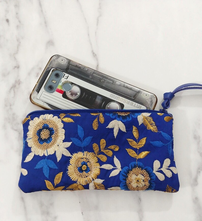 Op de afbeelding: Een koningsblauwe geborduurde clutch met een polsbandje, versierd met gouden, witte en blauwe bloemmotieven. Een smartphone met een cassettebandje hoesje ligt erin, een modieus accessoire.