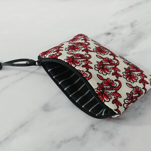 Op de afbeelding: Een kleine, open ritspouch met een bloemenpatroon. De pouch heeft een witte achtergrond met rode bloemmotieven. De binnenvoering is zwart-wit gestreept. Een zwarte rits en trekker maken het ontwerp compleet.