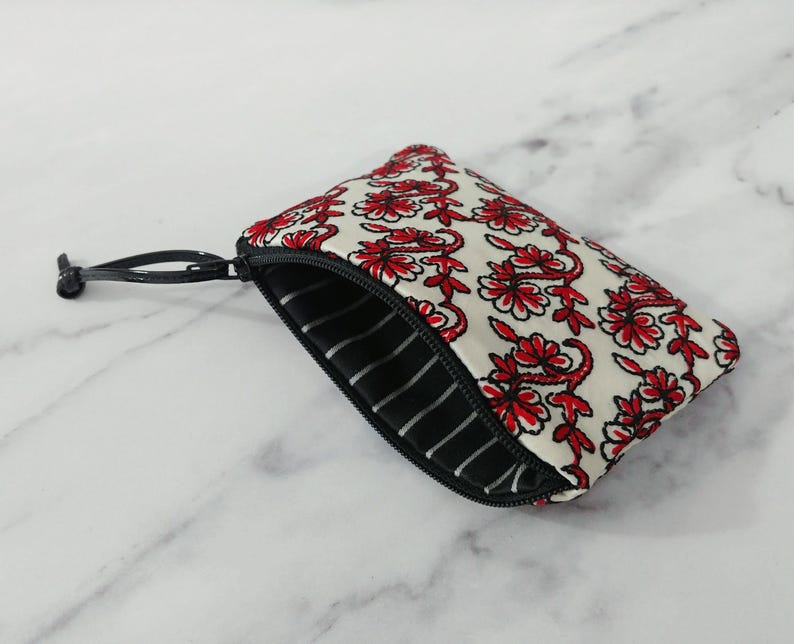 Op de afbeelding: Een kleine, open ritspouch met een bloemenpatroon. De pouch heeft een witte achtergrond met rode bloemmotieven. De binnenvoering is zwart-wit gestreept. Een zwarte rits en trekker maken het ontwerp compleet.