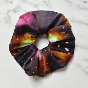 May include: A scrunchie with a galaxy print. The fabric features a blend of colors including pink, orange, green, and black, with white speckles resembling stars. The scrunchie is a hair accessory.