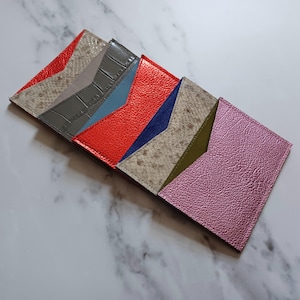May include: A collection of colorful leather cardholders in various shades, including red, gray, blue, and pink. Each cardholder features a unique texture and design, showcasing a variety of materials and patterns. The cardholders are arranged in a diagonal row.