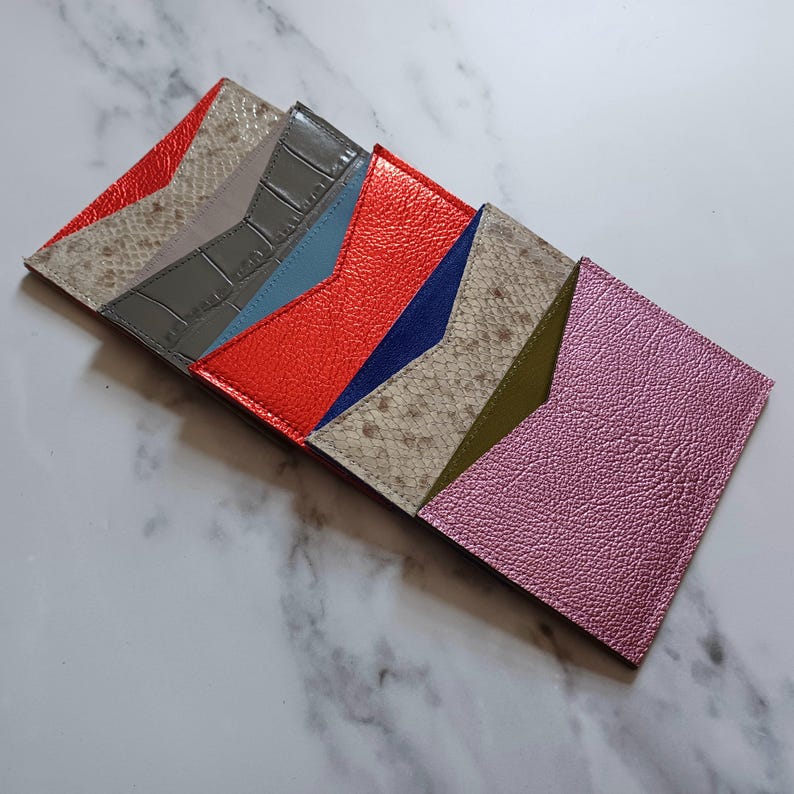May include: A collection of colorful leather cardholders in various shades, including red, gray, blue, and pink. Each cardholder features a unique texture and design, showcasing a variety of materials and patterns. The cardholders are arranged in a diagonal row.