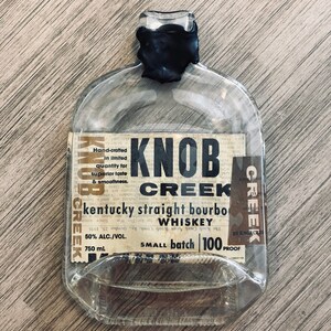 May include: An empty clear glass Knob Creek Kentucky straight bourbon whiskey bottle with a black wax seal on the neck. The label features black text on a cream background, stating 50% alc./vol., 750 mL, and 100 proof, with a small batch designation.