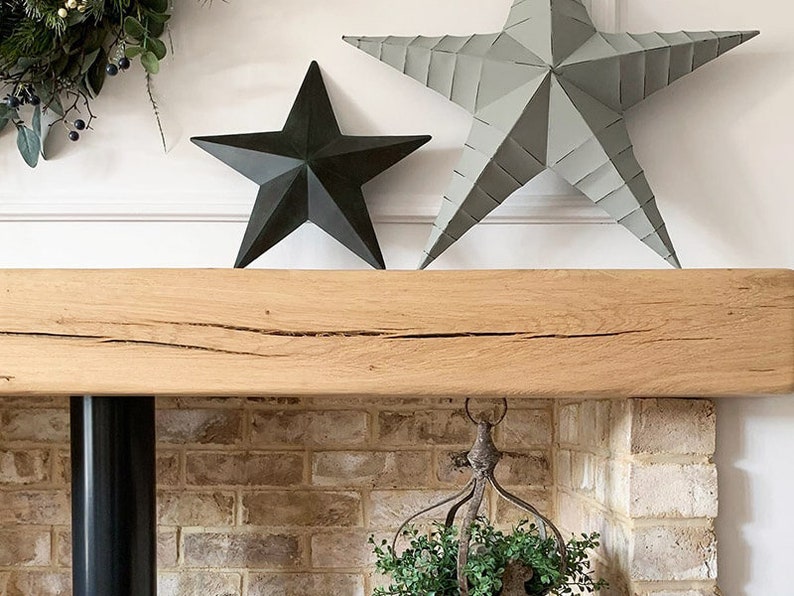May include: A rustic wooden mantel with a distressed finish, featuring two metal star decorations in gray and black. The mantel is mounted above a brick fireplace.