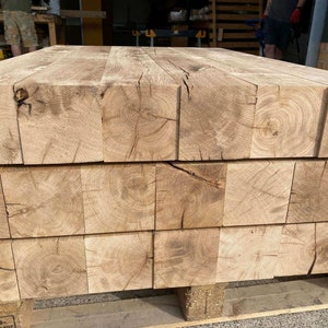 May include: Stacked wooden beams made of light brown oak with visible knots and grain patterns. The beams are arranged in a rectangular shape and are sitting on a wooden pallet.