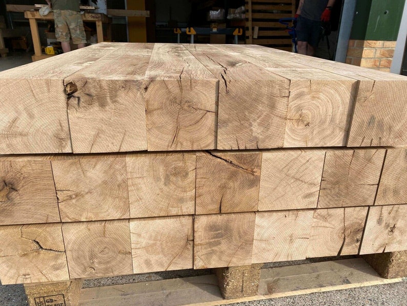 May include: Stacked wooden beams made of light brown oak with visible knots and grain patterns. The beams are arranged in a rectangular shape and are sitting on a wooden pallet.