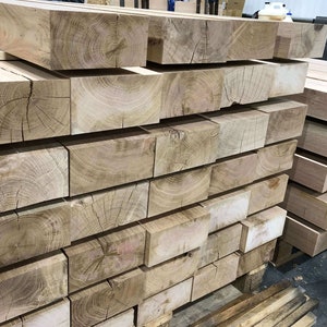 May include: A stack of large, rectangular wooden beams with visible grain patterns. The beams are light brown in color and have a rough, unfinished texture.