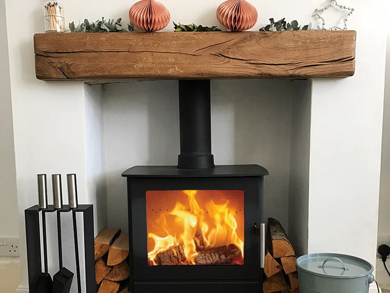 May include: A black wood-burning stove with a roaring fire inside. The stove is set into a white fireplace with a wooden mantel above. The mantel is decorated with two pink paper lanterns and greenery.