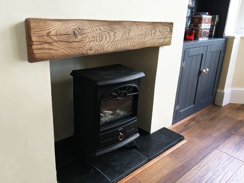 May include: A black electric fireplace with a realistic flame effect sits in a white fireplace surround with a rustic wooden mantel. The fireplace is in a room with dark wood floors and a dark wood cabinet with doors.