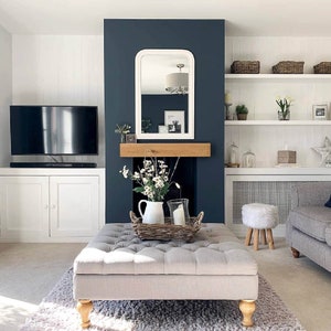 May include: A living room with a white and gray sofa, a white ottoman with tufted upholstery, a white fireplace with a dark blue mantle, and a white shelf unit with baskets and decorative items.