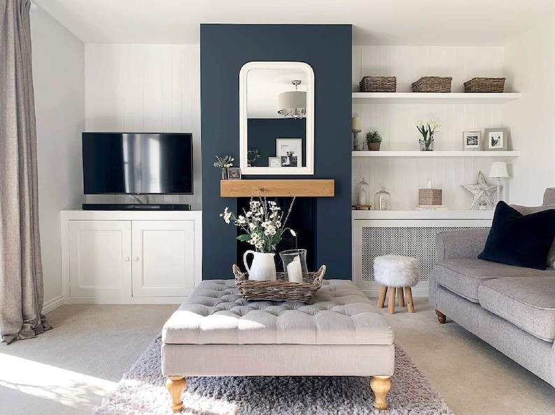 May include: A living room with a white and gray sofa, a white ottoman with tufted upholstery, a white fireplace with a dark blue mantle, and a white shelf unit with baskets and decorative items.