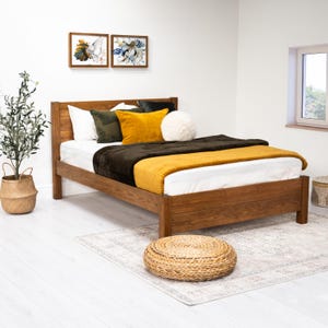 May include: A wooden bed frame with a white mattress, brown blanket, and yellow throw. The bed is styled with pillows in various colours and textures. The bed is in a room with white walls and a rug on the floor.