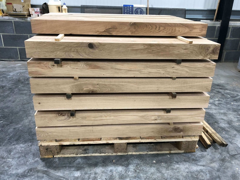 May include: A stack of six wooden planks on a pallet. The planks are light brown and have a rough texture. The pallet is made of wood and has a brown stain.