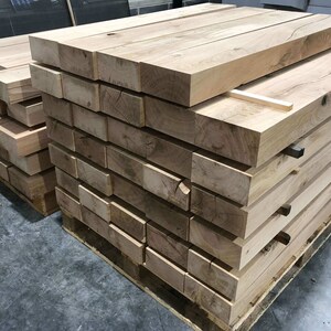 May include: A stack of wooden planks, likely for woodworking projects, are stacked on a pallet. The planks are light brown in color and have a rough texture.