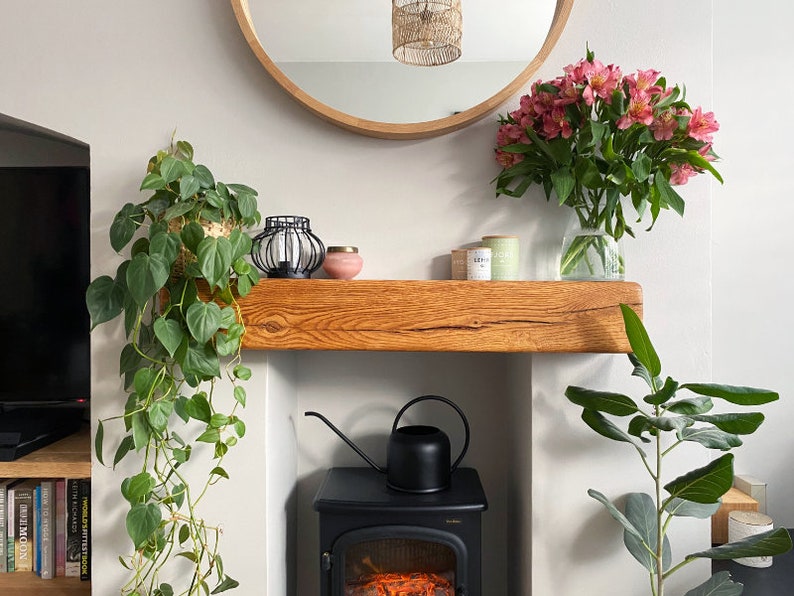May include: A wooden mantel shelf with a potted plant, a vase of pink flowers, and a black watering can. The shelf is mounted above a black fireplace with a glass door.