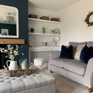 May include: A gray sofa with two dark blue throw pillows and a faux fur throw pillow. The sofa is in a living room with a white fireplace mantel, a white shelf unit, and a white ottoman with a wicker tray on top.