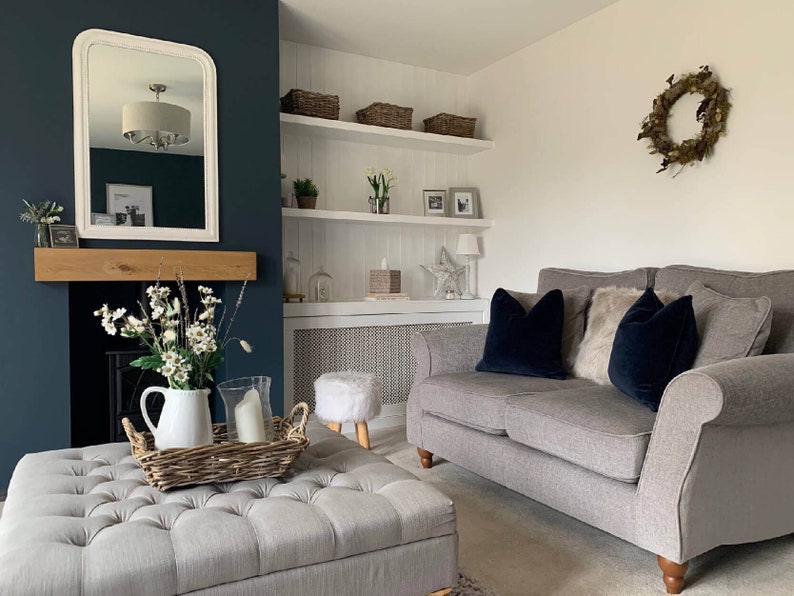 May include: A gray sofa with two dark blue throw pillows and a faux fur throw pillow. The sofa is in a living room with a white fireplace mantel, a white shelf unit, and a white ottoman with a wicker tray on top.