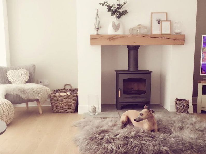 May include: A living room with a fireplace, a wood-burning stove, a grey armchair with a white heart-shaped pillow, a wicker basket, a grey fluffy rug, and a small dog lying on the rug.